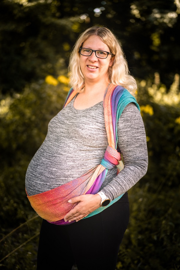 Frau die im 9. Monat schwanger ist, trägt ein buntes Tragetuch. Das Tragetuch ist Regenbogenfarben. Sie hat das Tuch so gebunden, dass es ihren großen Bauch stützt. 