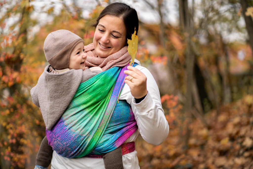 Mama trägt ihr einjähriges Kind in einem bunten Tragetuch. Im Hintergrund sieht man einen herbstlichen Wald