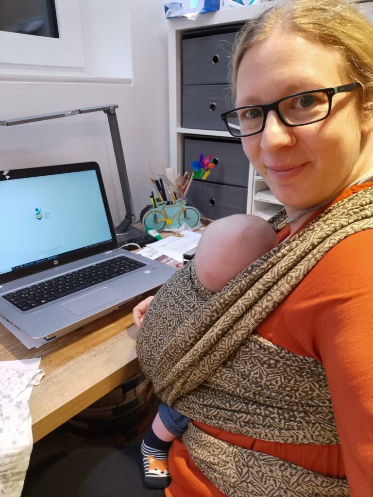 Mutter arbeitet am Schreibtisch mit Baby in Trage. Beispiel für Alltag nach Trageberatung