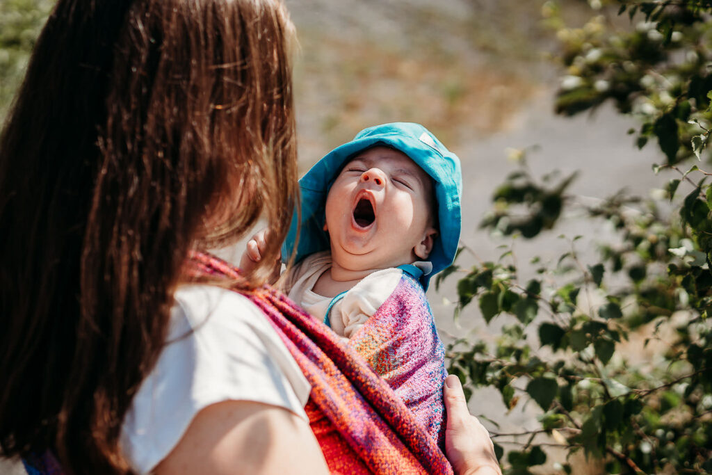 Mutter trägt gähnendes Baby im bunten Tragetuch. 